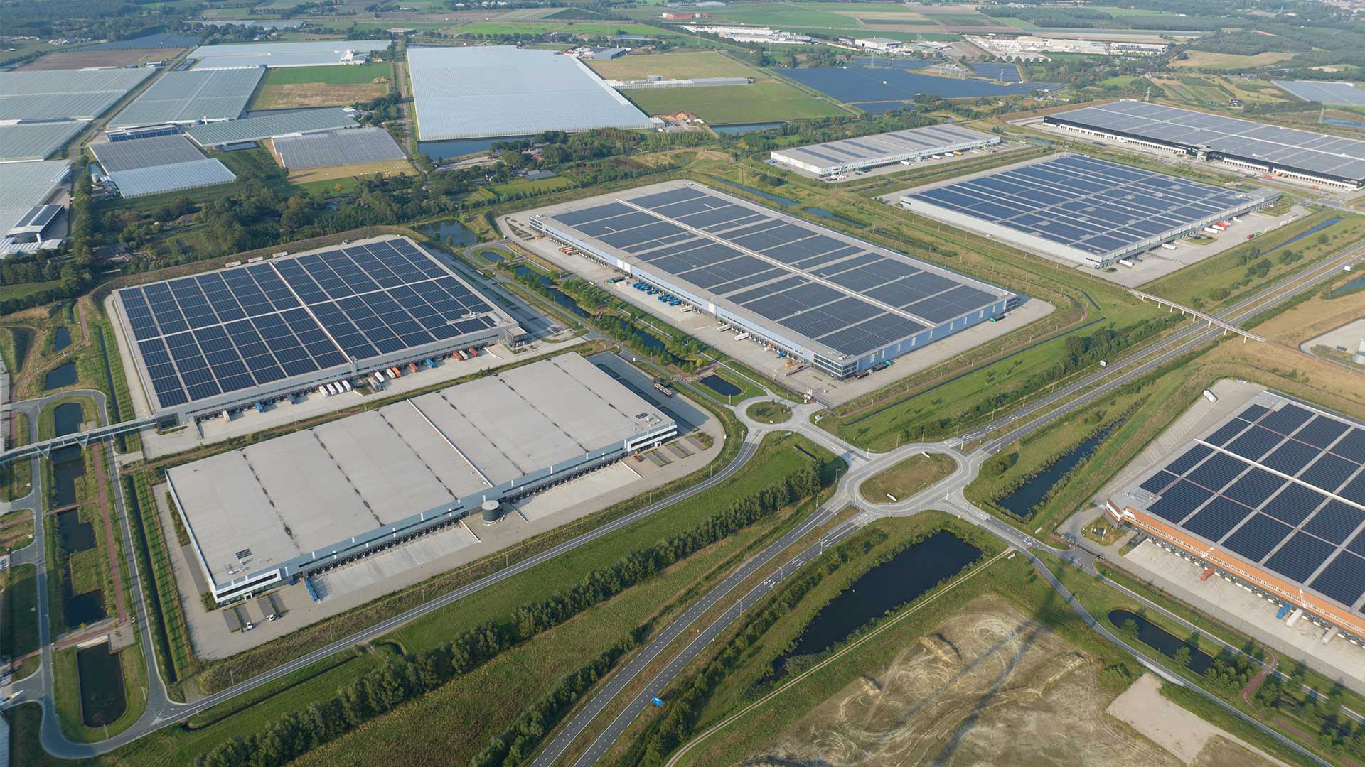 Aerial view of large logistics park with distribution centers and warehouse roofs covered with solar panels, connected by roads and waterways.