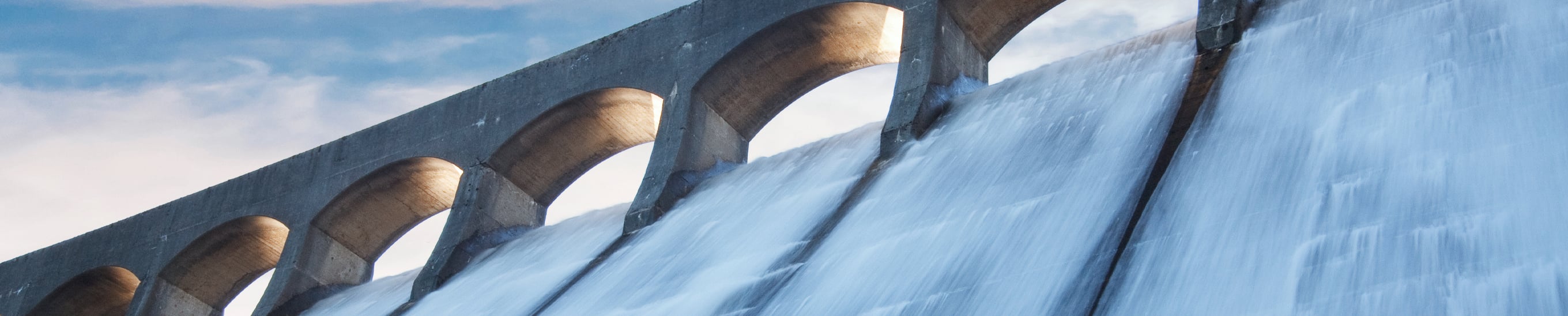 Close-up of water cascading down a large concrete hydroelectric dam spillway with arched supports against a blue sky