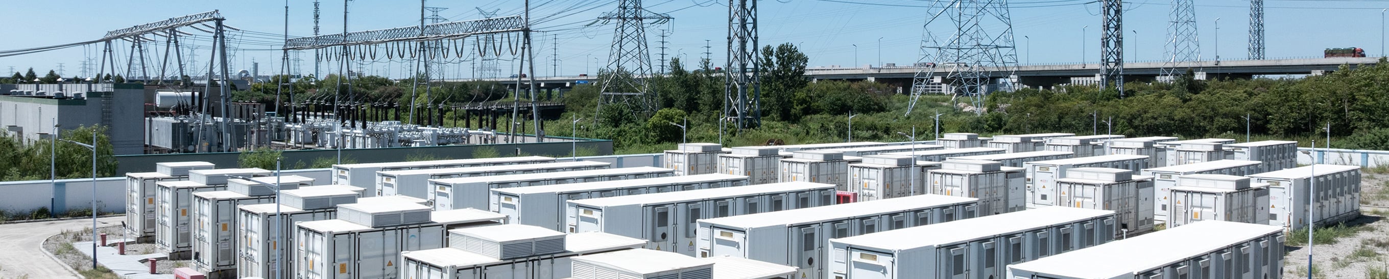 Panoramic view of a utility-scale battery energy storage facility with rows of white containerized units adjacent to a high-voltage electrical substation and transmission towers
