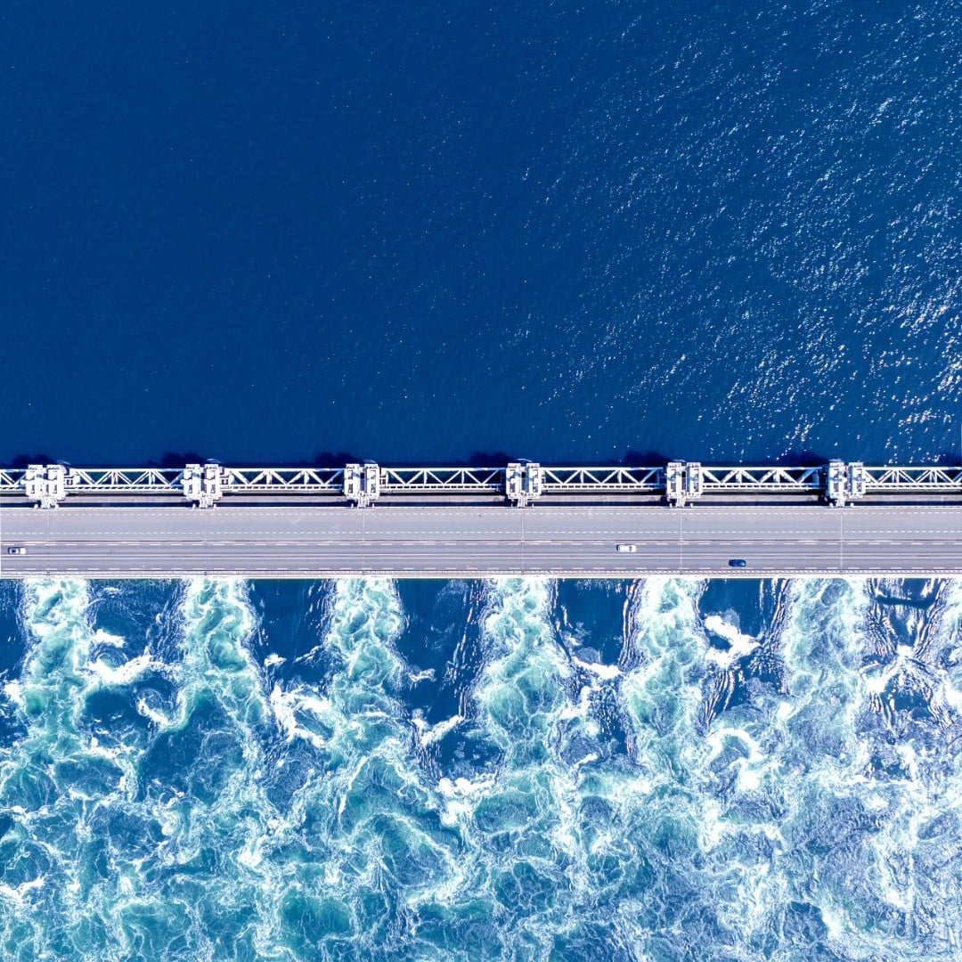 Aerial view of a storm surge barrier bridge separating calm deep water from turbulent churning water, illustrating CBRE's Q3 2026 Macro House View theme of economic resilience through persistent geopolitical and energy shocks.