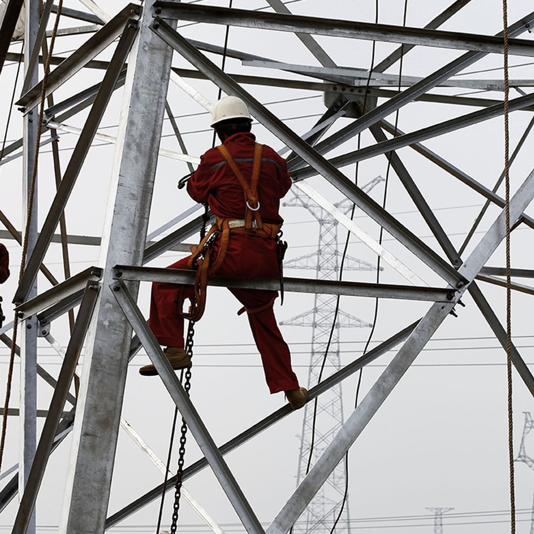 Utility worker in red safety gear climbing a steel power transmission tower