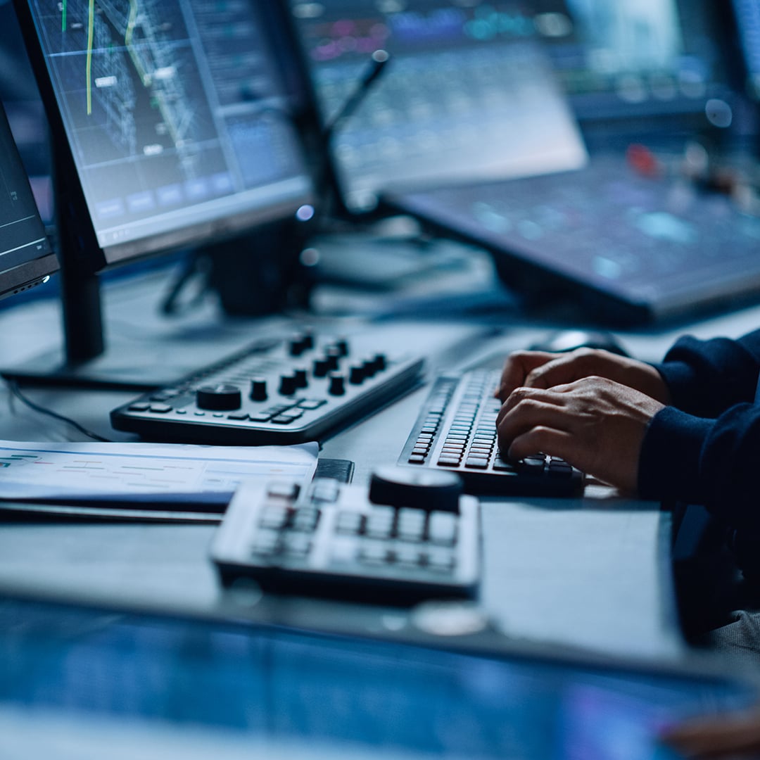 Person typing at a workstation with multiple monitors displaying data analytics and system dashboards.