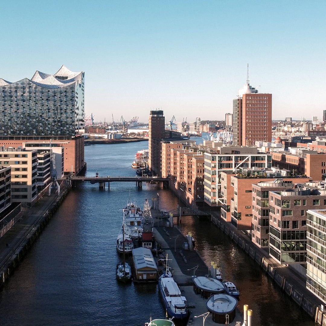 Aerial view of Hamburg's HafenCity district featuring the iconic Elbphilharmonie concert hall, a tree-lined canal with moored boats, brick warehouse buildings, and the port skyline under a clear blue sky
