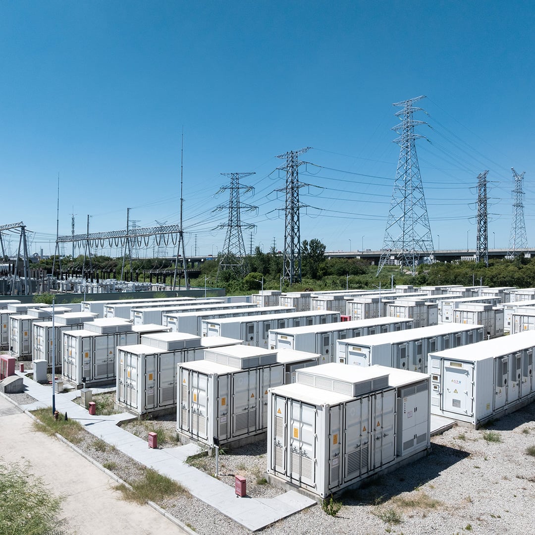 Aerial view of a large-scale battery energy storage facility with rows of white containerized battery units adjacent to a high-voltage electrical substation and transmission towers under a clear blue sky