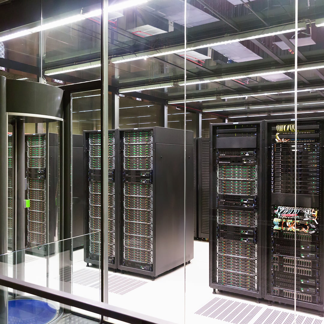Interior of a modern data center featuring rows of black server racks with green indicator lights behind glass security partitions and overhead LED lighting