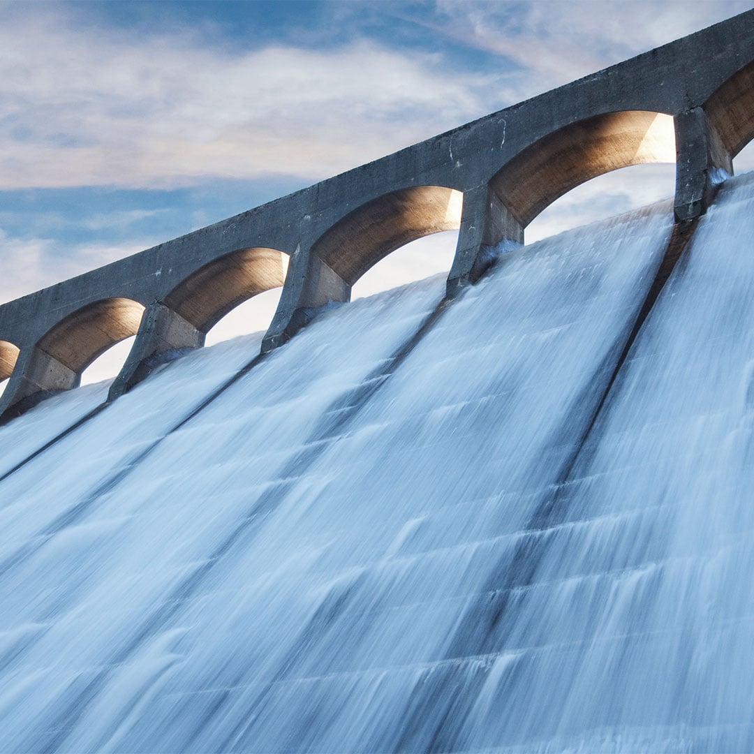 Water cascading down the spillway of a large concrete dam, with arched supports visible along the top against a partly cloudy sky.