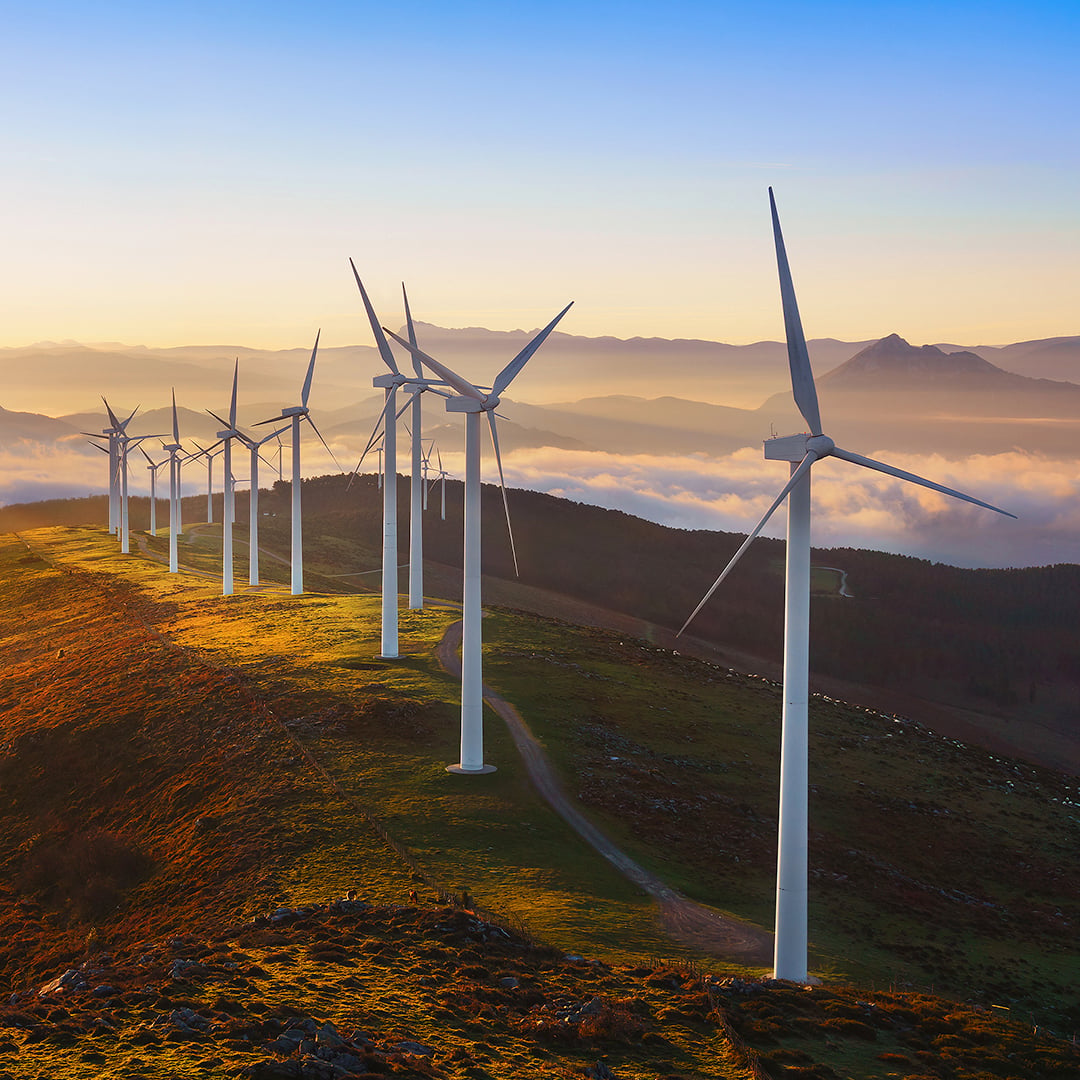 Row of white wind turbines along a green hillside at sunrise, with mountains and low clouds in the background, representing renewable energy infrastructure investment.