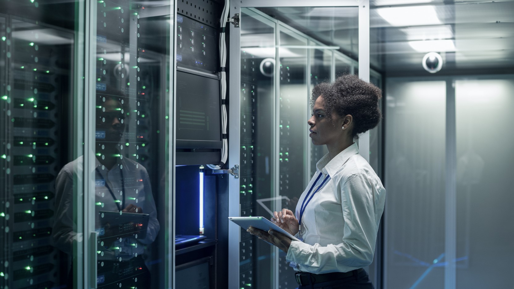 A data center technician holding a tablet inspects an open server rack in a secured facility with glowing green indicator lights and glass-enclosed server rows