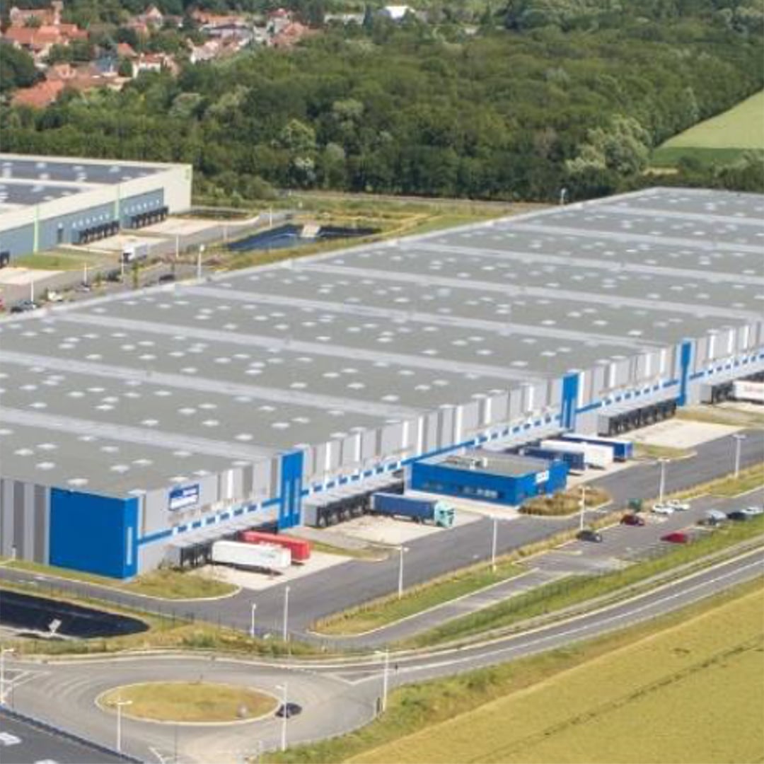 Aerial view of a large modern industrial warehouse complex with loading docks, trucks, and surrounding greenery near a suburban area