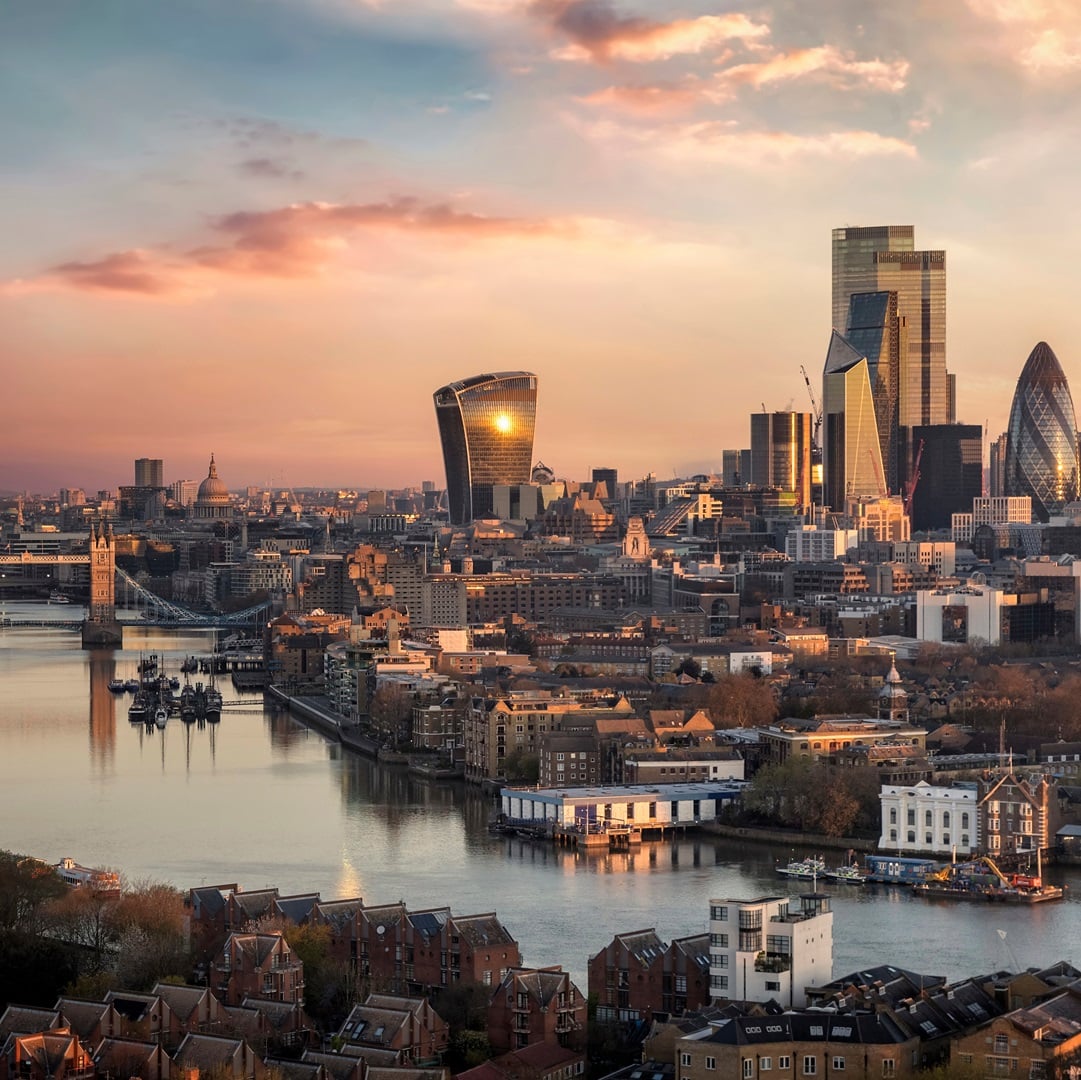 Aerial view of the London skyline along the River Thames featuring office towers, Tower Bridge, and the City of London financial district at sunset.
