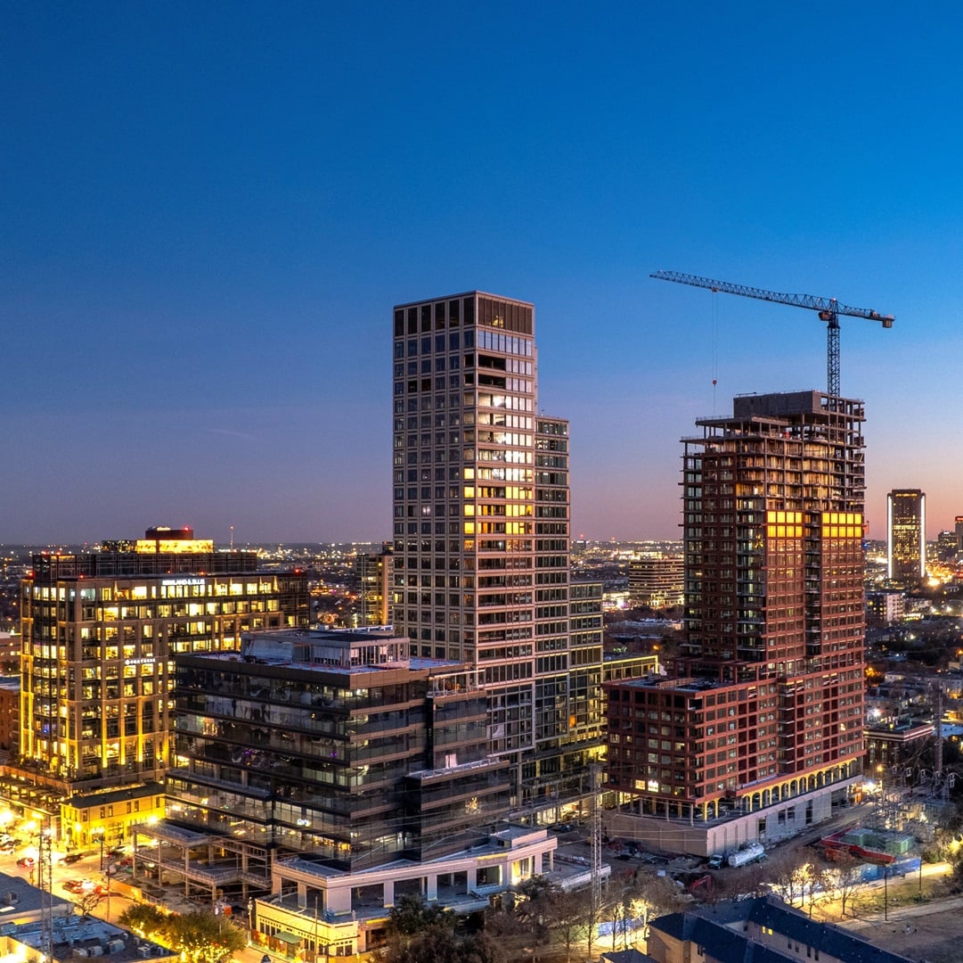 Aerial view of modern high-rise commercial buildings in a downtown skyline at dusk, with illuminated offices and a tower under construction.