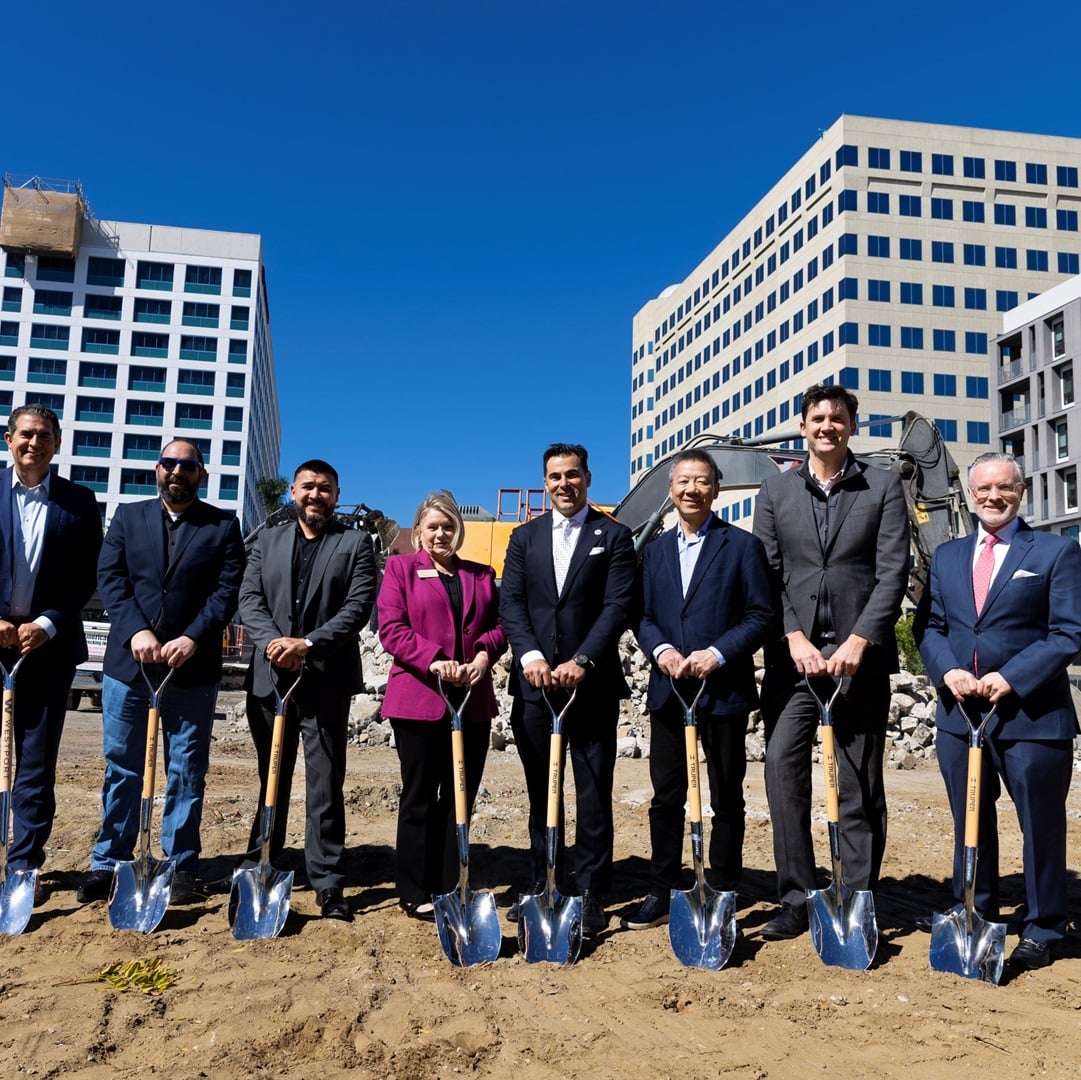 Business leaders participate in a ceremonial groundbreaking for a new commercial office development with construction equipment and office buildings in the background.