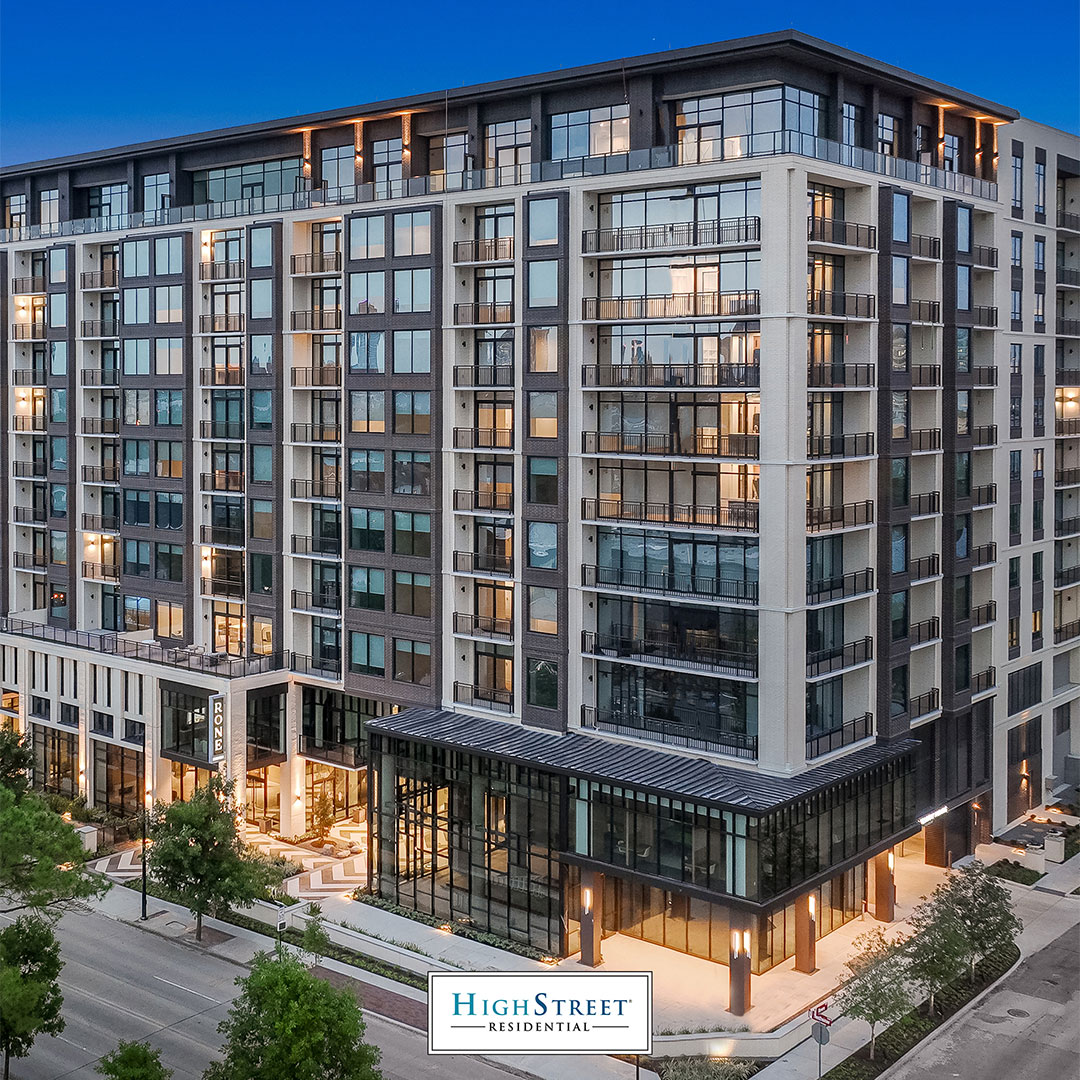 Modern mid-rise apartment building with glass balconies and ground-floor retail on a city street at dusk.