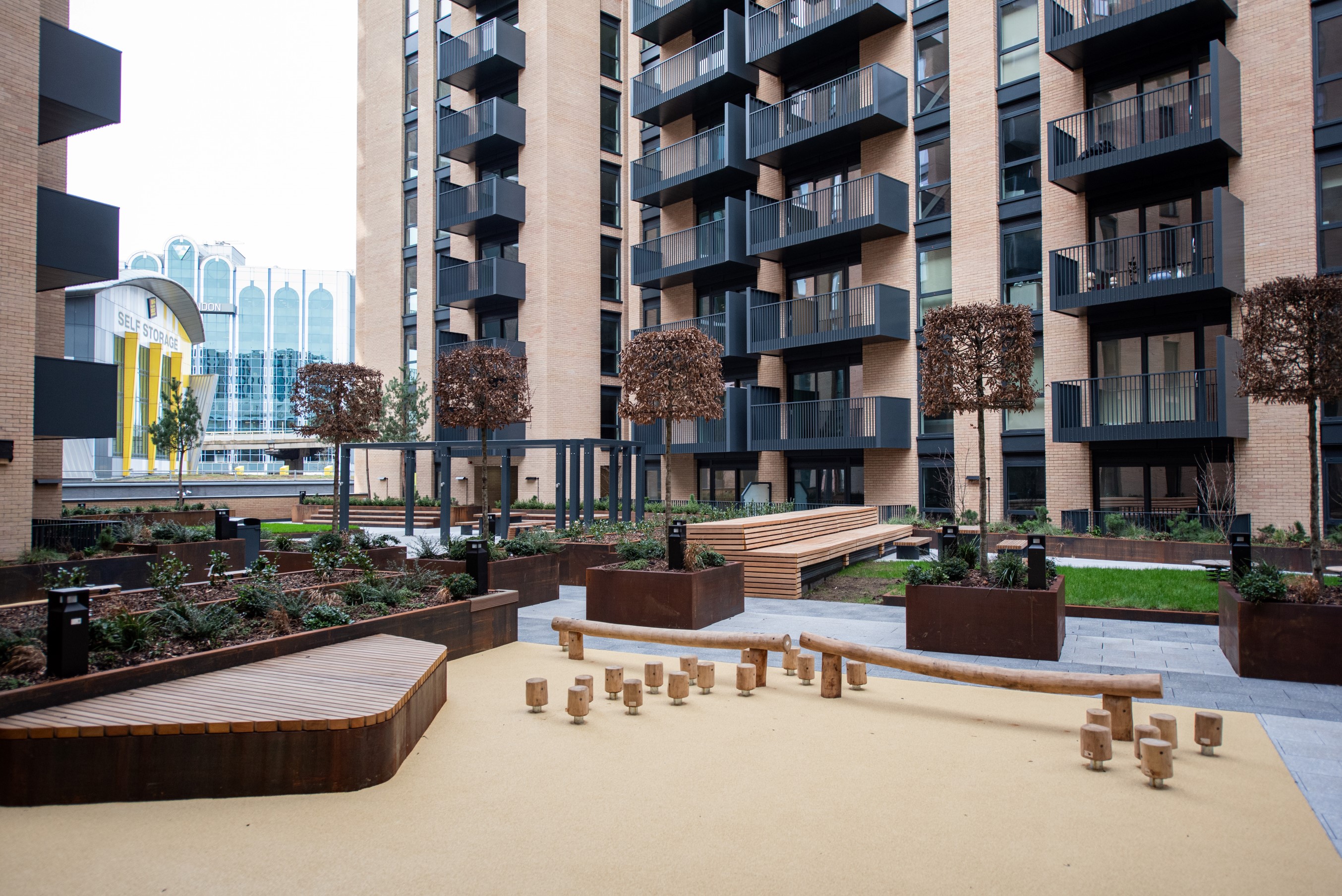 Modern apartment complex with landscaped courtyard, seating areas, play elements, and private balconies on residential buildings.