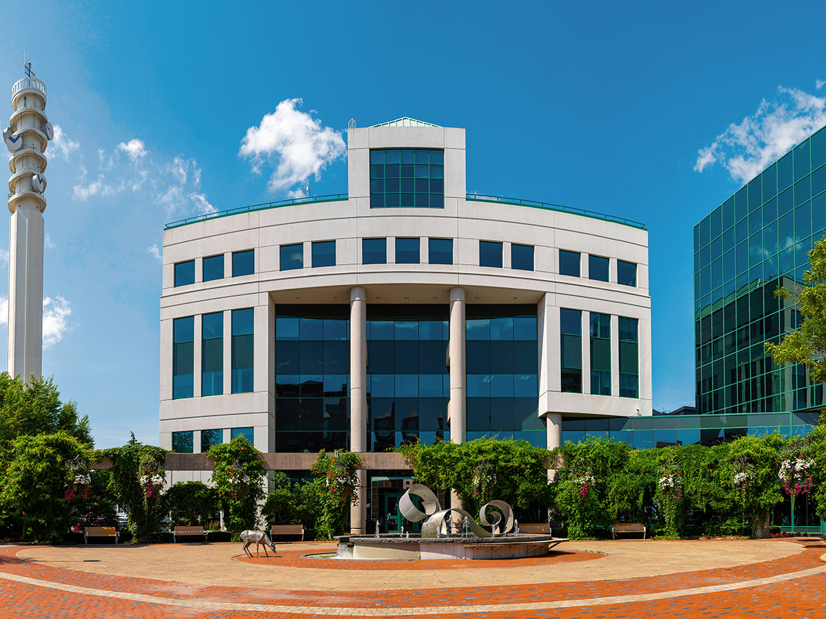 A scenic view of the Moncton skyline featuring a modern commercial building under a clear blue sky, with large reflective windows, an adjacent white tower, and a public space with benches and greenery