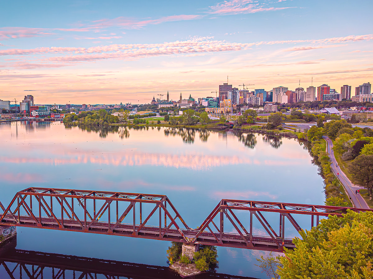 Aerial view of Ottawa skyline at sunrise with a historic railway bridge over a calm river, reflecting the cityscape and colorful sky. The image showcases the blend of urban development and natural beauty 