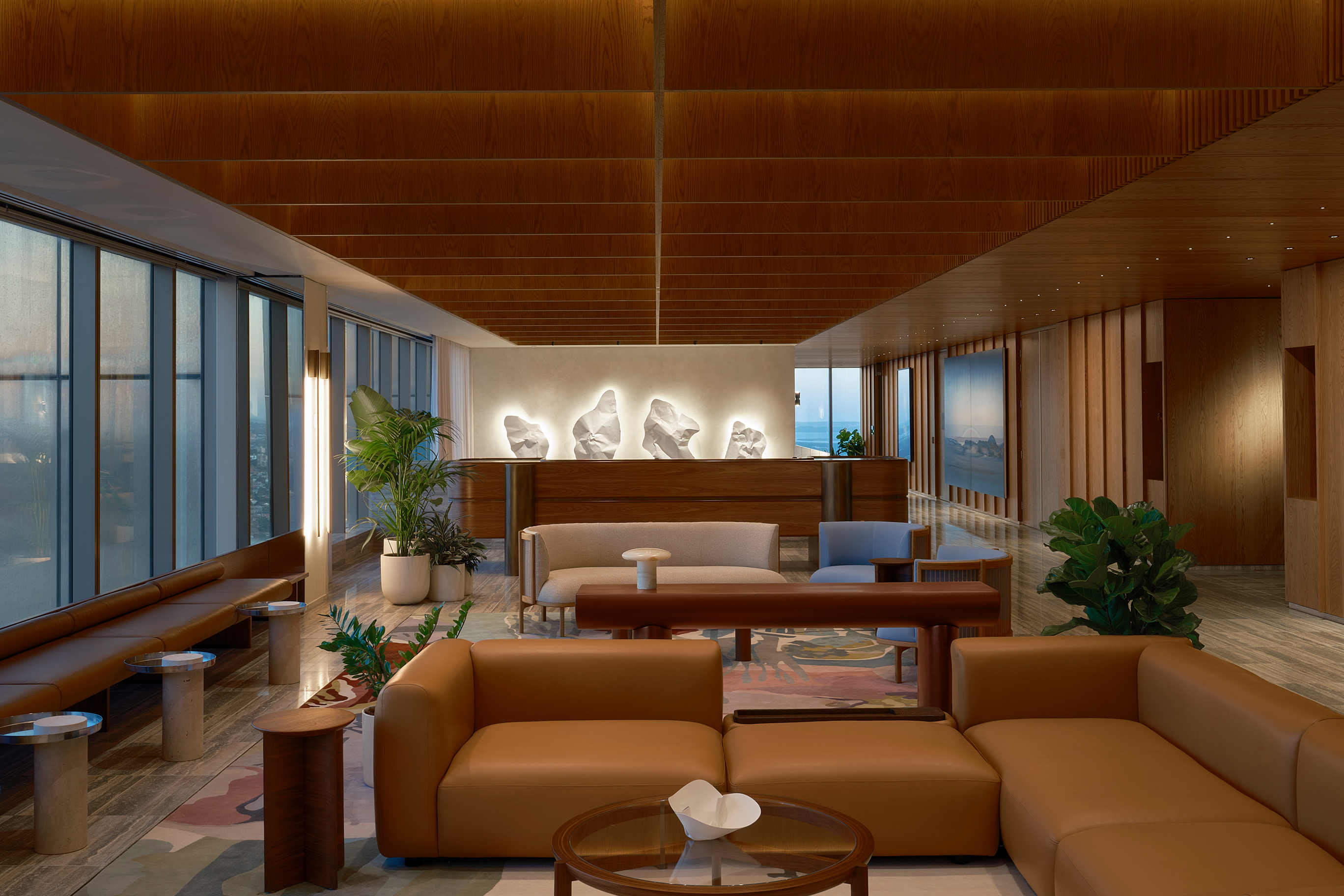 This image depicts a luxurious, modern lounge area with large windows offering a view of the outside, featuring comfortable seating arrangements in earthy tones and sleek wooden finishes throughout.