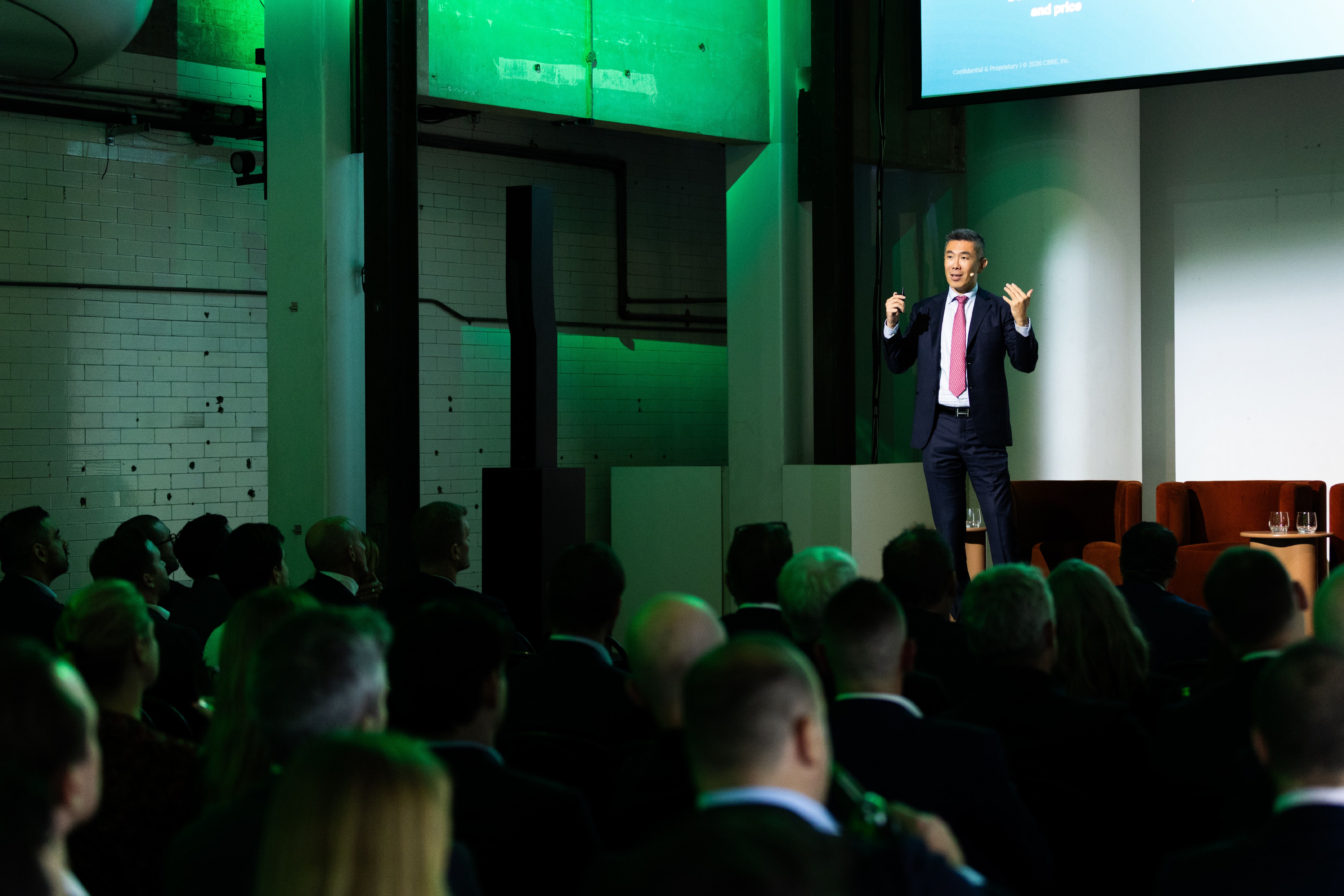A man in a suit gestures while speaking to a large audience in an industrial event space.