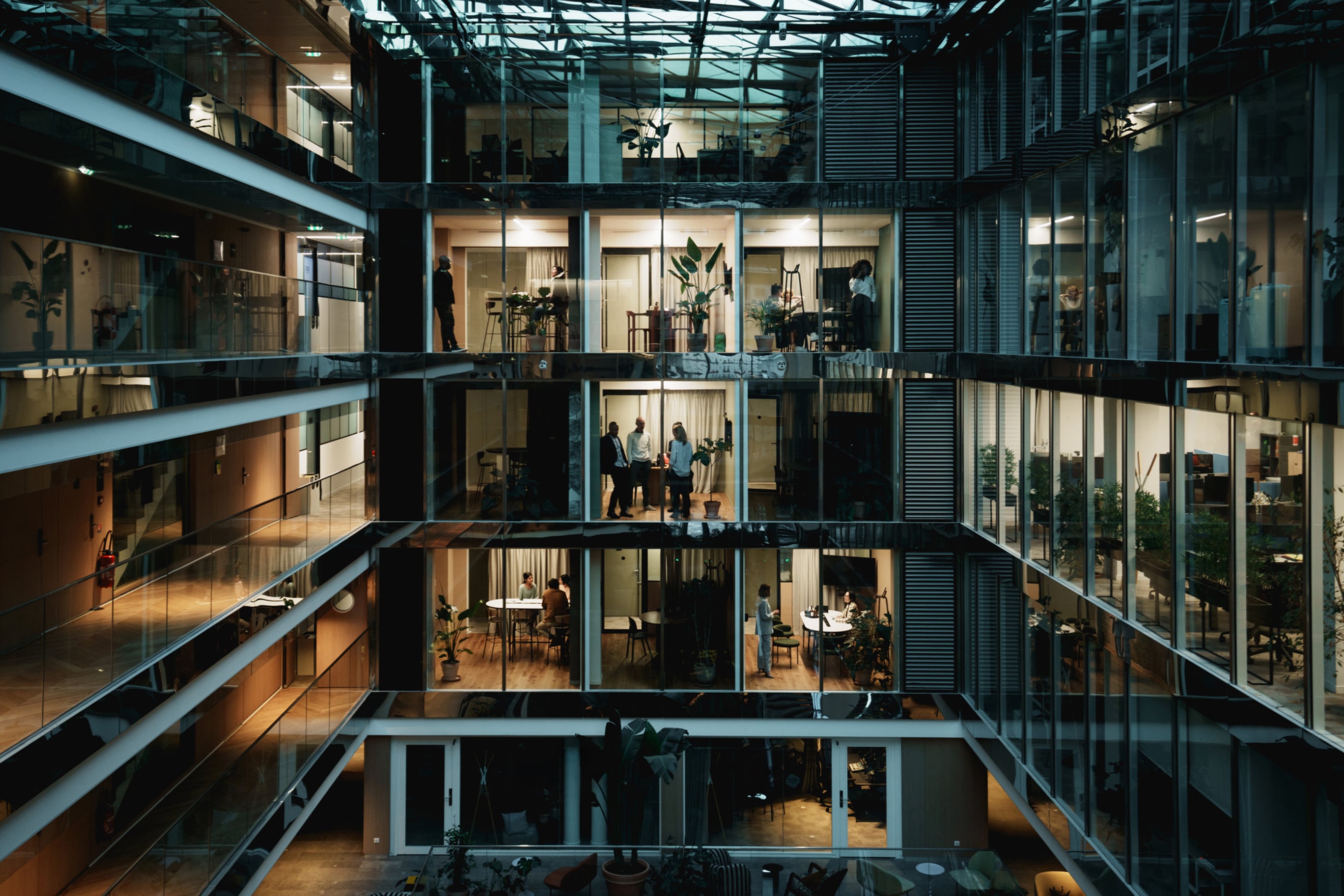 A multi-storey office building with glass-walled rooms reveals various employees working and socialising in a modern, open-plan atrium.