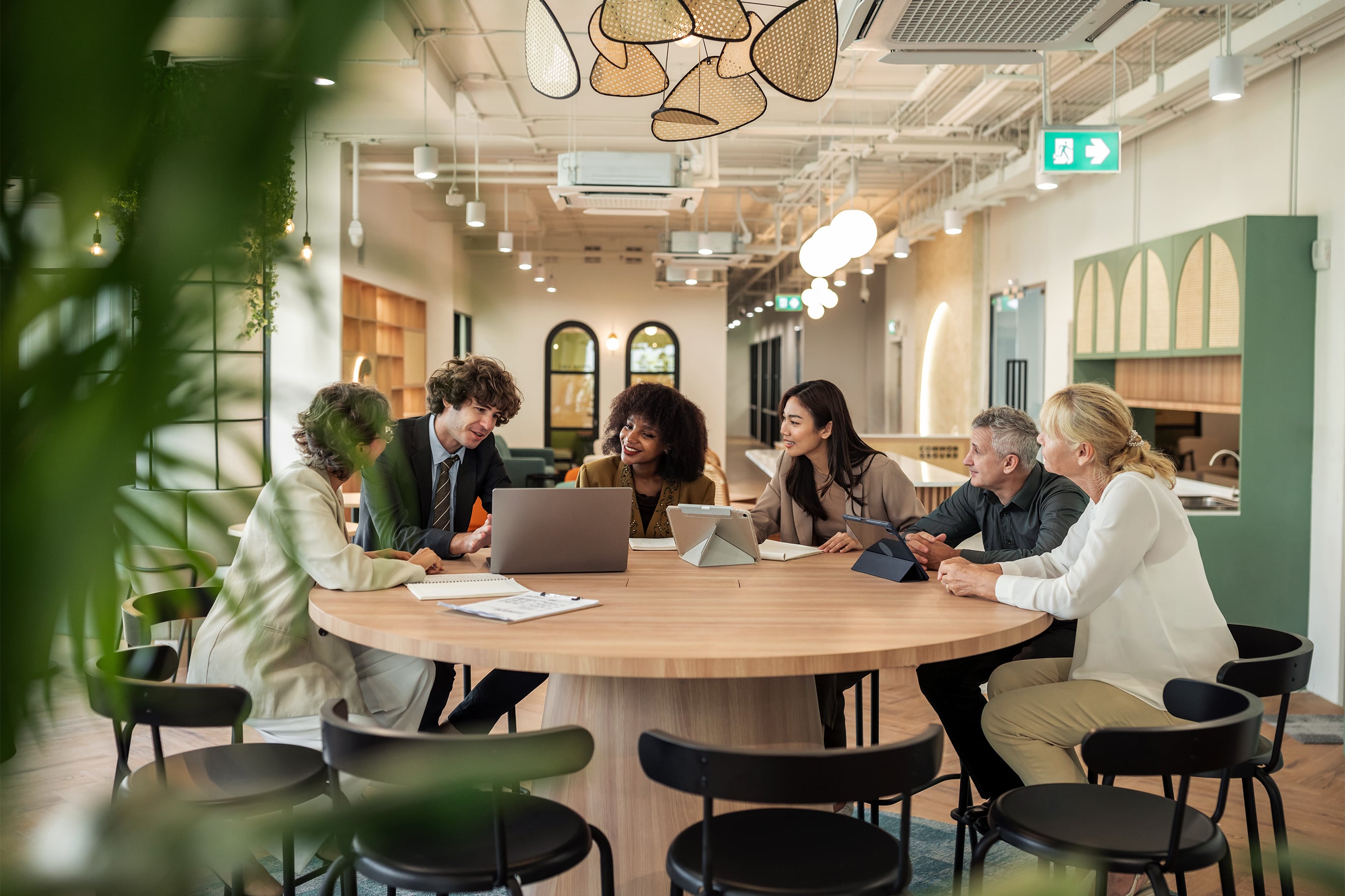 A diverse group of professional colleagues sit around a large circular table in a brightly lit, modern office space, collaborating on a project using laptops and tablets.