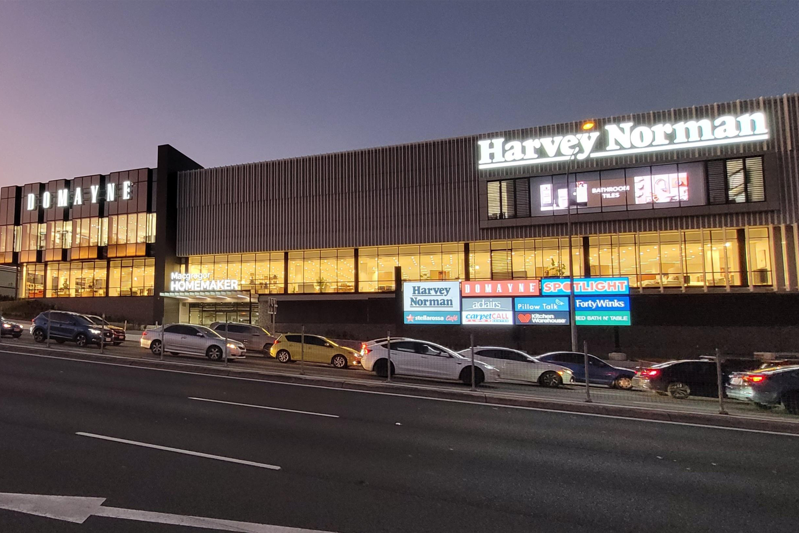 An evening view of a large shopping complex, "Domayne" and "Harvey Norman" signs prominently displayed, with several cars along the street.