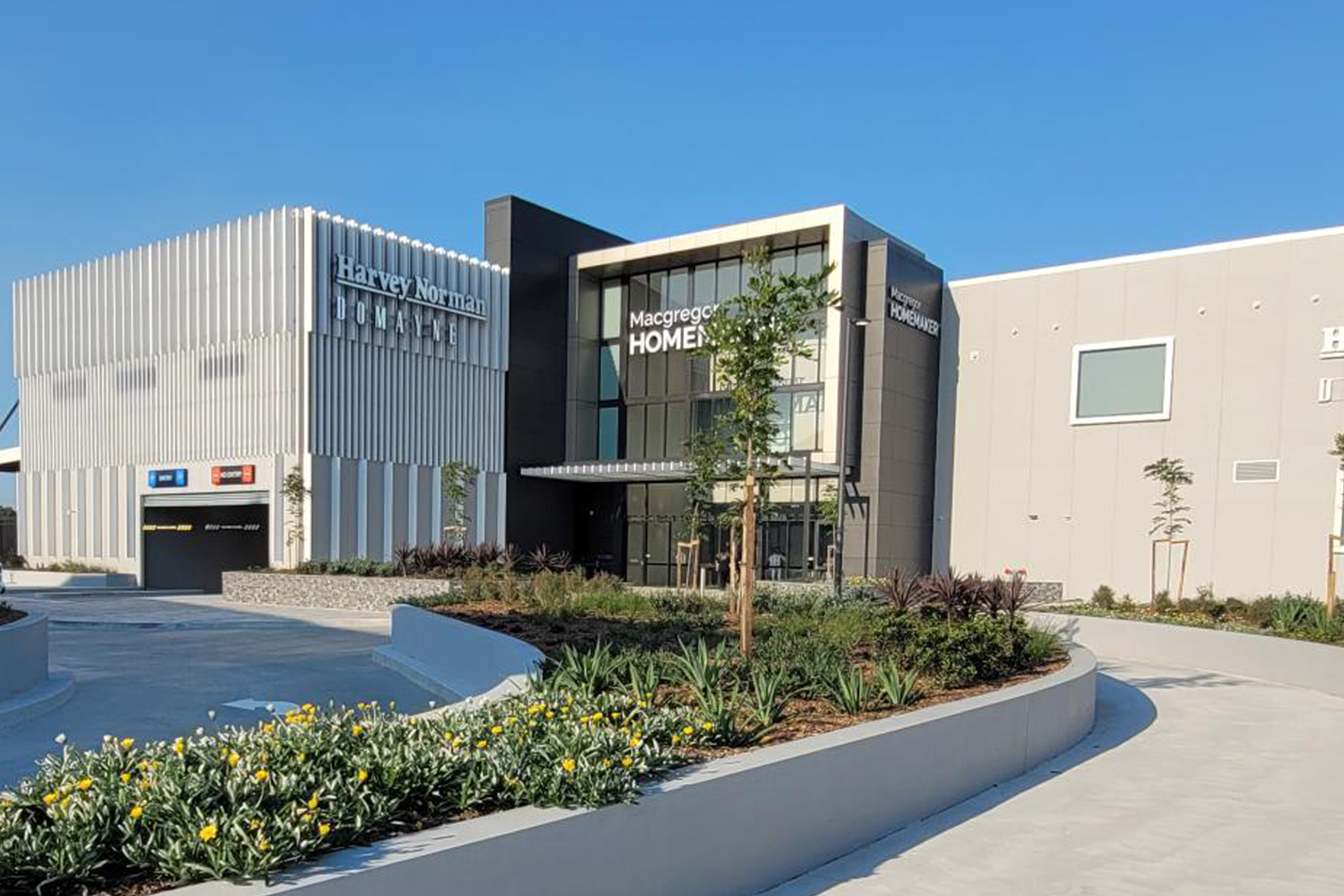 A modern retail complex with a white corrugated Harvey Norman building and a dark grey Macgregor Homemaker building, both surrounded by landscaped gardens and a clear blue sky.