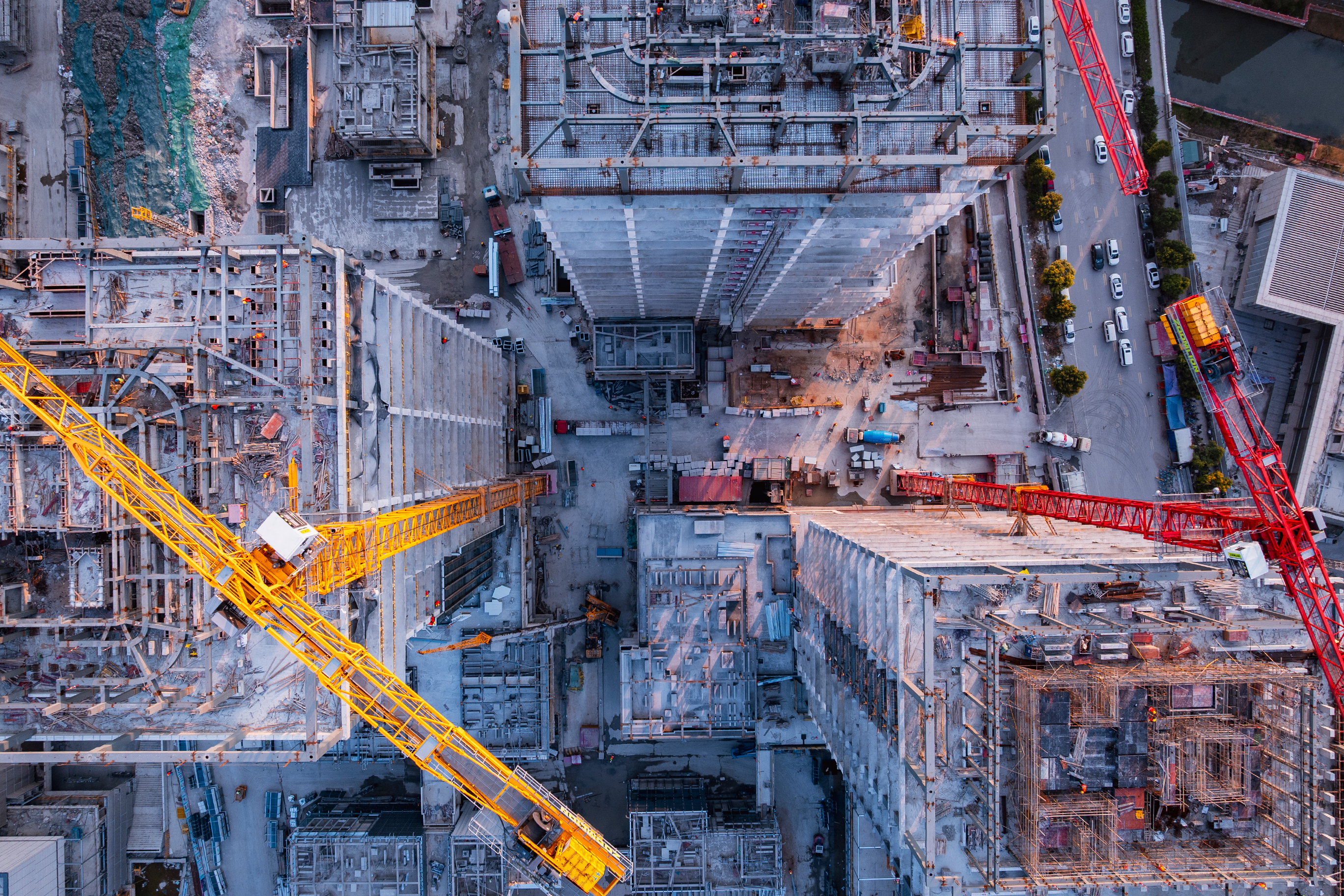 An aerial view from above a large construction site shows multiple buildings in various stages of completion, with several yellow and red cranes towering over the structures.