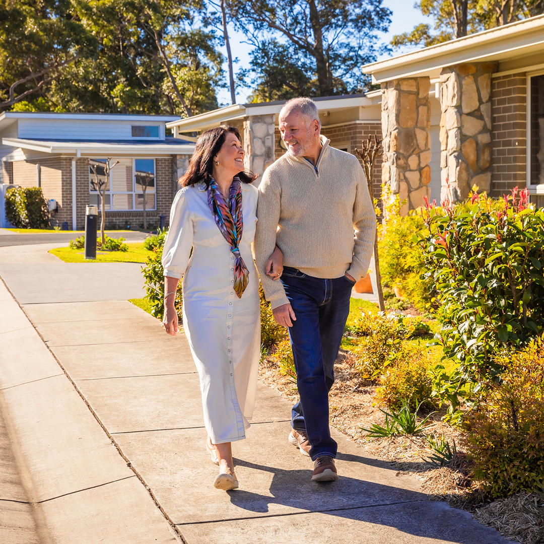 A smiling older couple strolls arm-in-arm along a sunny suburban footpath outside their home.