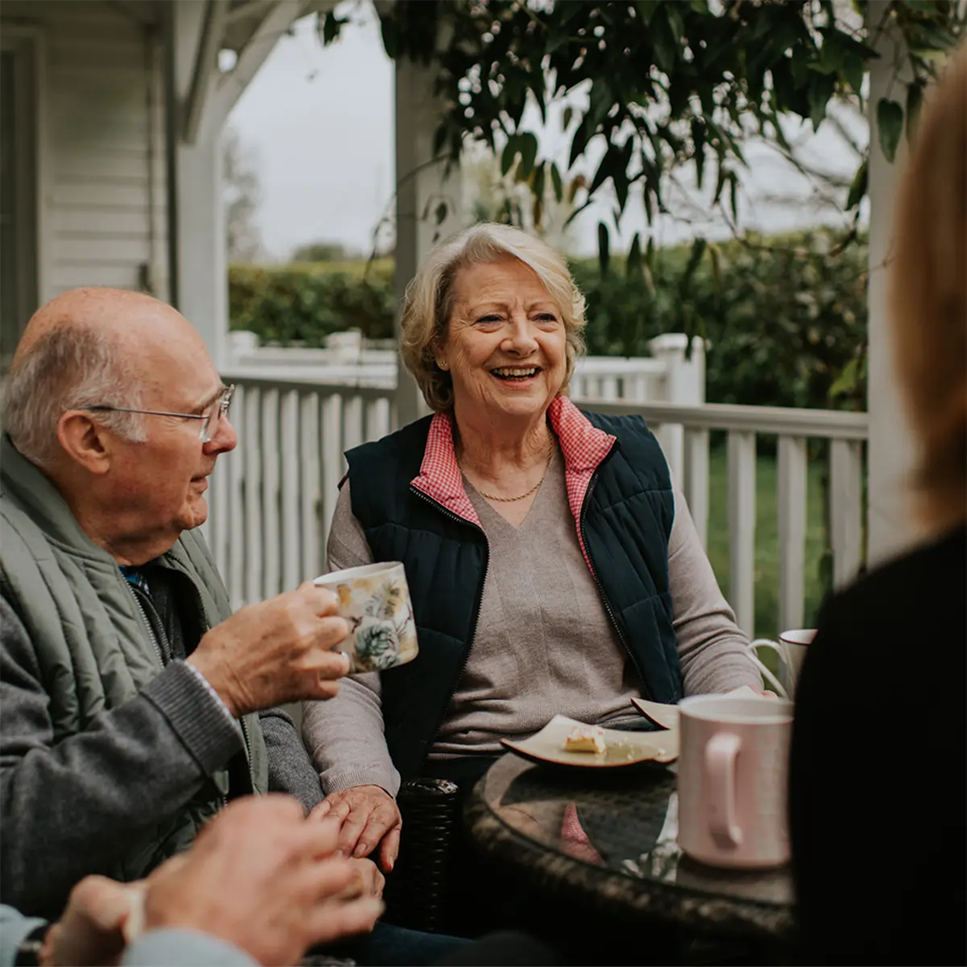A group of older adults sit together on a verandah, enjoying tea and conversation in a relaxed outdoor setting.