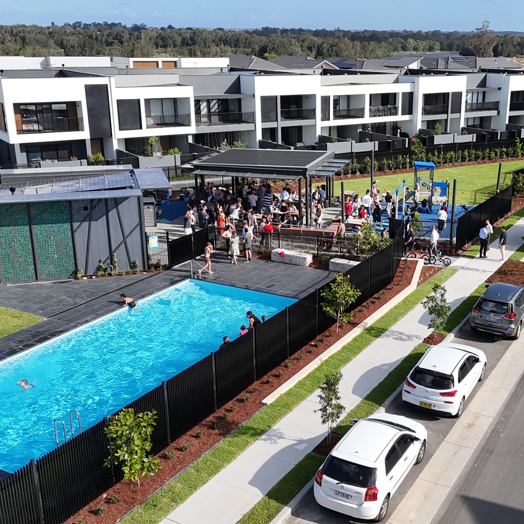 An aerial view shows a vibrant community gathering around a swimming pool, shaded outdoor area, and playground, surrounded by modern white and grey townhouses with parked cars along the street.