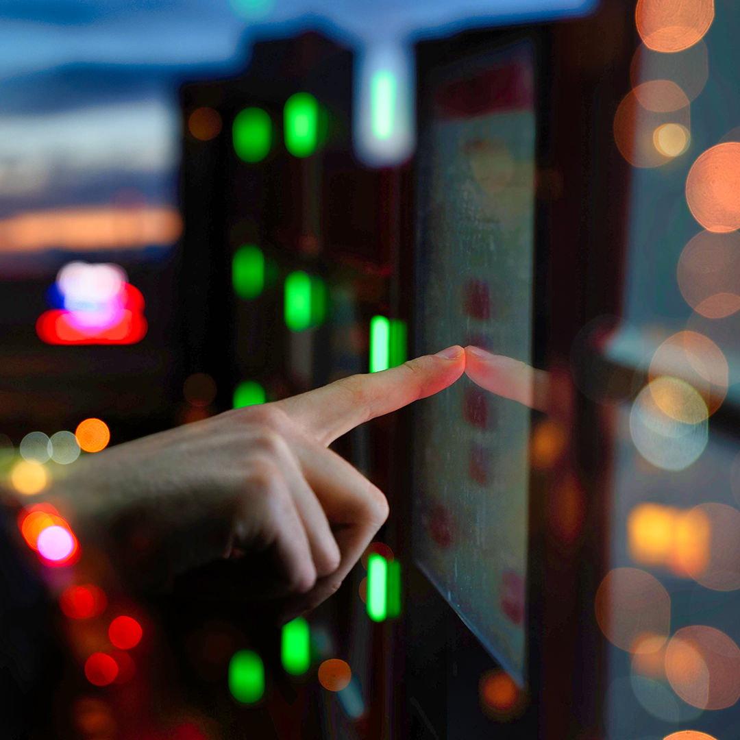A person's hand with an extended index finger touches a reflective screen, surrounded by blurred colourful lights in a dark setting.