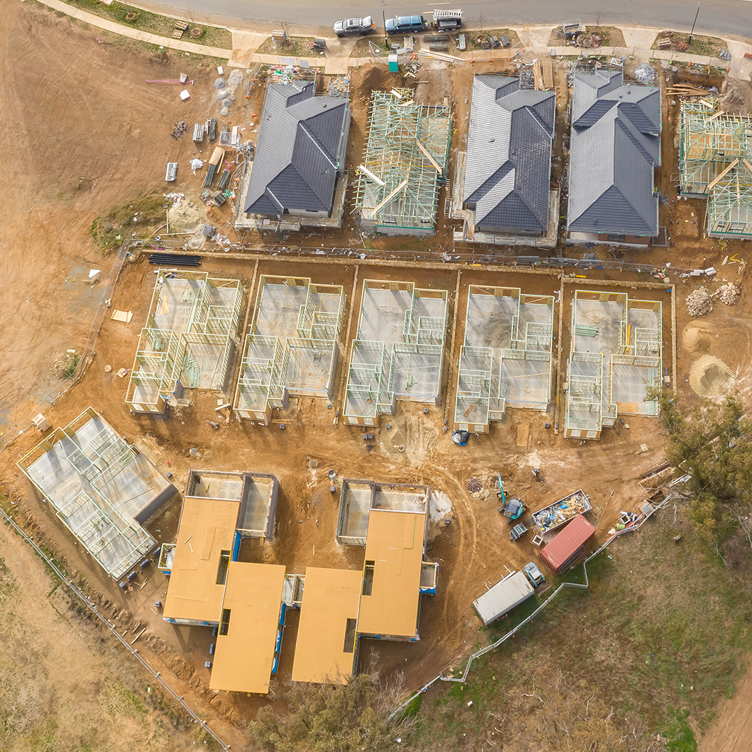An aerial view shows a housing estate development in various stages of construction on a large, mostly dirt site.