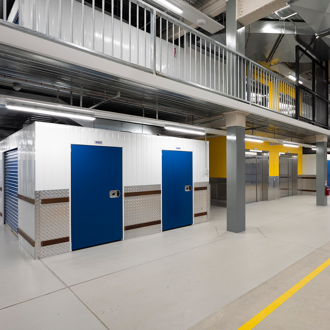 A modern self-storage facility features white corrugated metal units with bright blue doors on the ground floor, beneath a mezzanine level with a metal railing and exposed ventilation ducts.