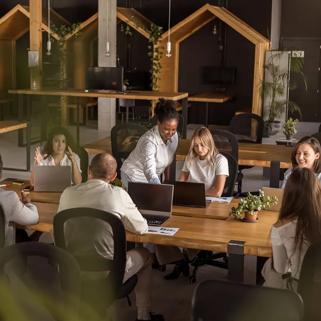 A diverse group of people are collaboratively working on laptops around a large wooden table in a modern office.