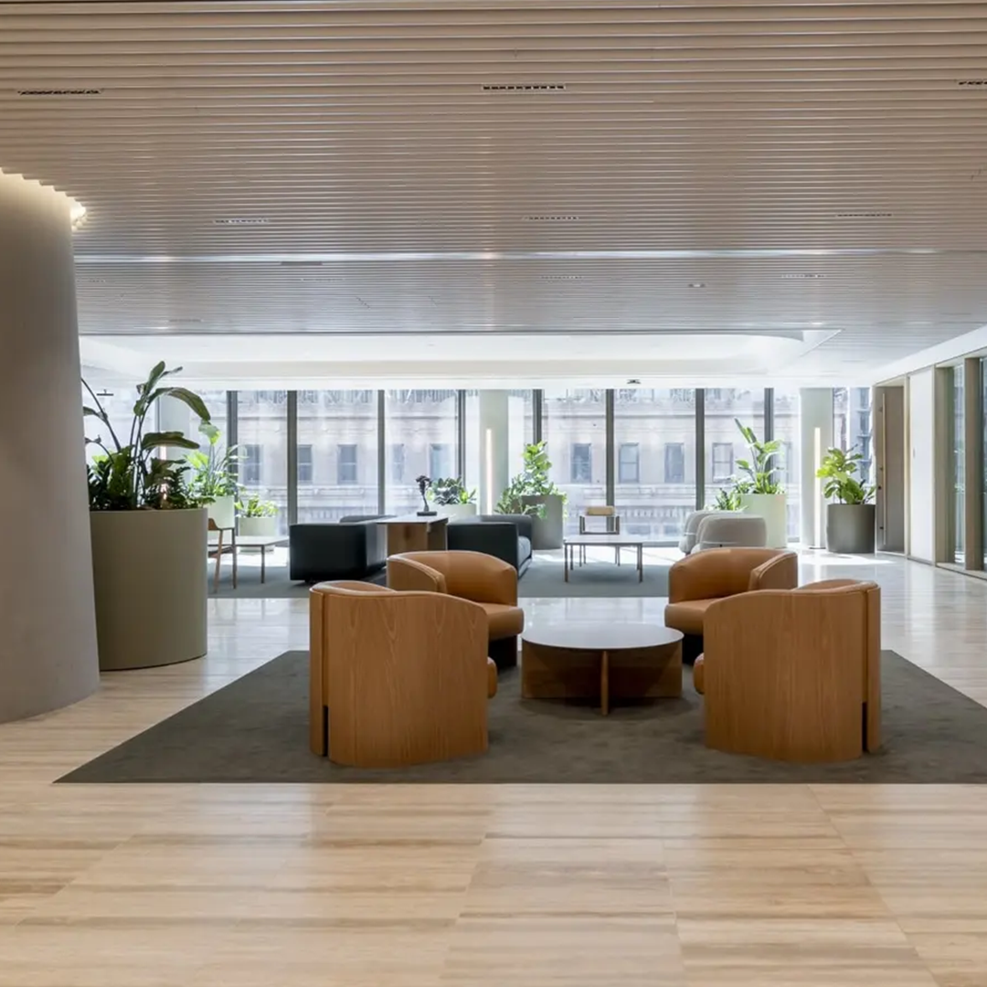 A modern, sunlit office lobby features light wooden armchairs around a coffee table on a dark rug, with large potted plants and city views through expansive windows.