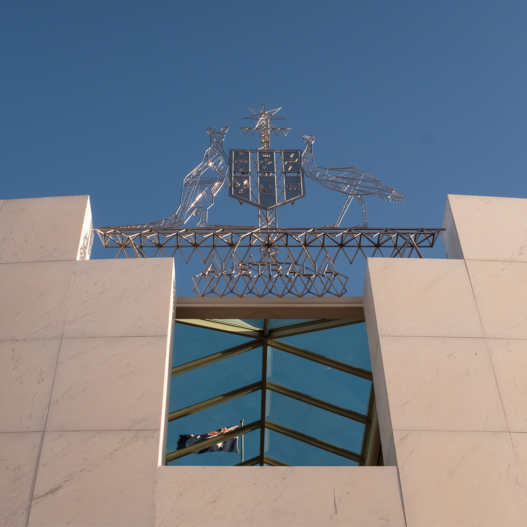 The Australian coat of arms, featuring a kangaroo and an emu, sits atop a modern building with a large glass window reflecting the Australian flag.