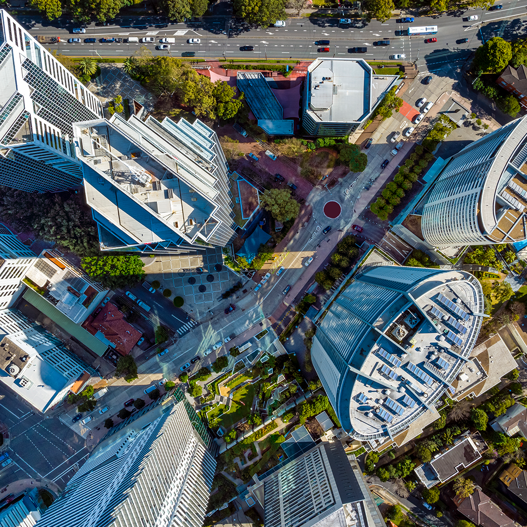 An aerial view captures modern high-rise buildings and roads, with a central roundabout and green spaces nestled amongst them, all bathed in bright sunlight.