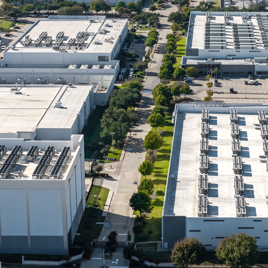 An aerial view shows several large, white industrial buildings with numerous HVAC units on their roofs, separated by a tree-lined road and green spaces.