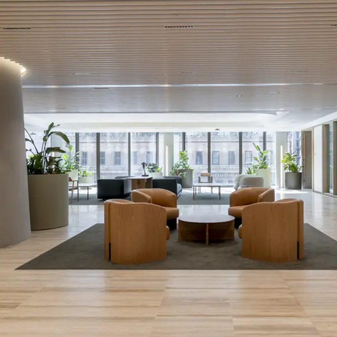 A bright, modern office waiting area features light wood flooring, several comfortable seating arrangements, and large windows overlooking a city street.