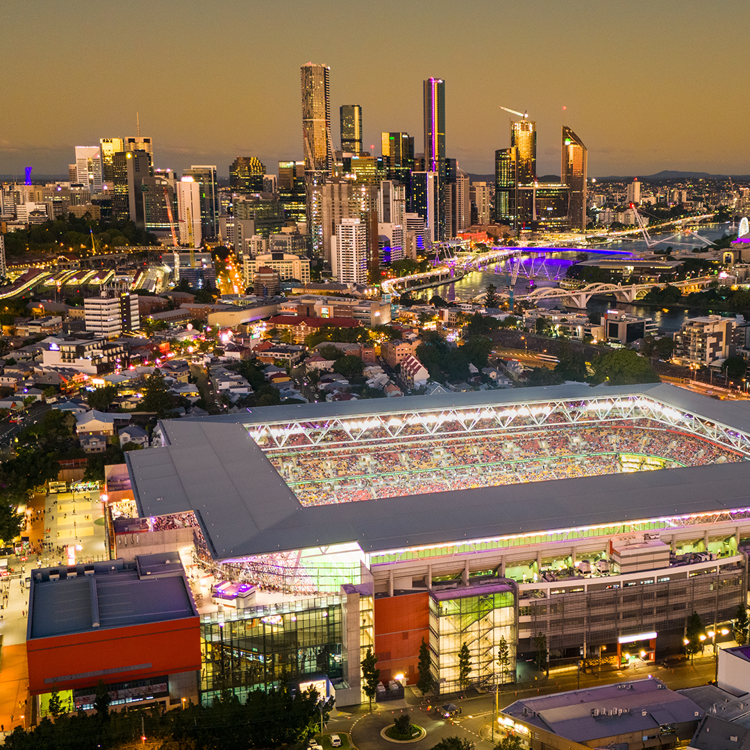 An aerial view at dusk shows Suncorp Stadium in Brisbane, Queensland, brightly lit and packed with spectators, against a backdrop of the city skyline and the Brisbane River.