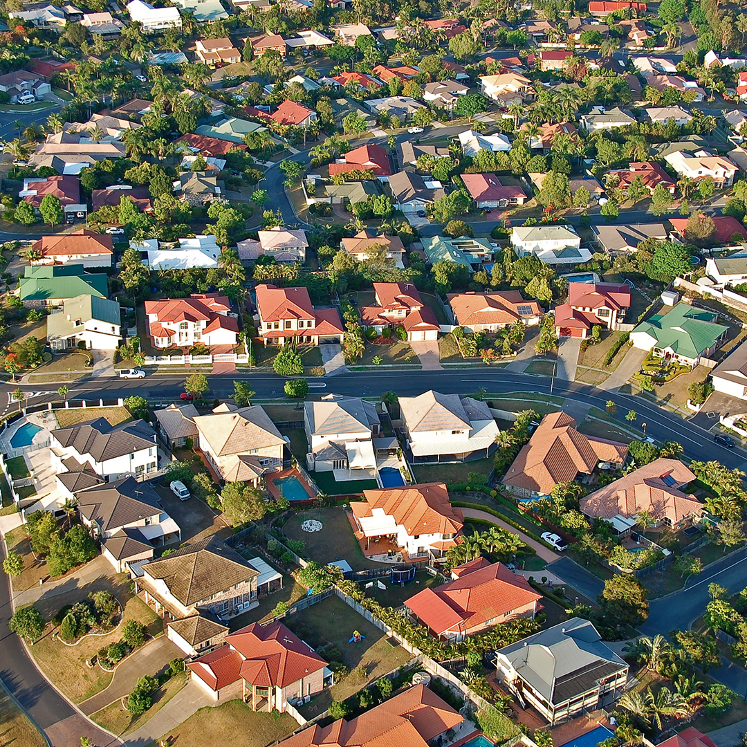 An aerial shot captures a sprawling Australian suburban neighbourhood with numerous houses featuring red and terracotta roofs, interspersed with lush green trees and winding roads.