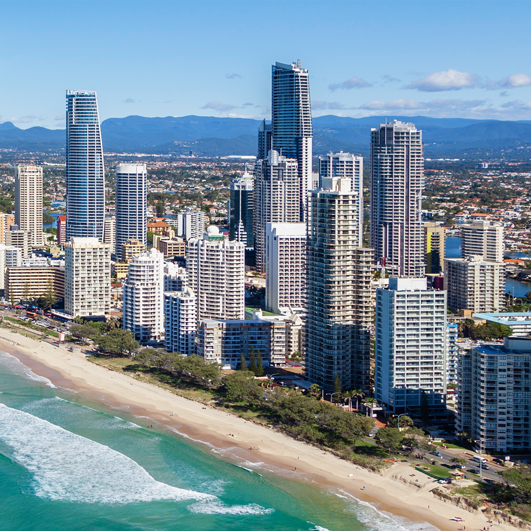 An aerial view shows the bustling Gold Coast skyline, with numerous high-rise buildings towering over a sandy beach and turquoise ocean, all set against a backdrop of distant mountains under a clear blue sky.