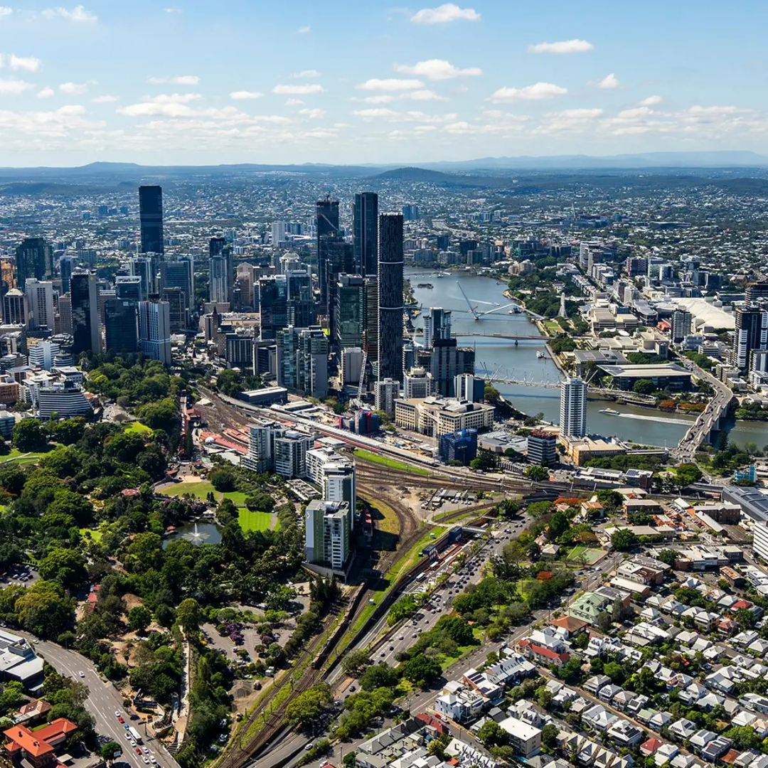 An aerial view captures the sprawling Brisbane city skyline, with its towering buildings, a winding river featuring several bridges, and lush green parks, all under a bright blue sky with scattered clouds.
