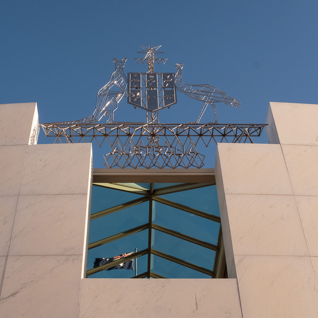 A grand, metallic Australian Coat of Arms stands prominently against a clear blue sky, with a reflection of the Australian flag visible in the windows below.