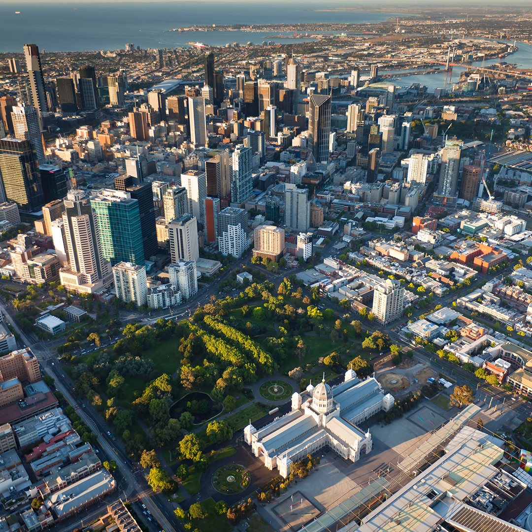 An aerial view of Melbourne shows the Royal Exhibition Building and Carlton Gardens surrounded by city skyscrapers, with the bay in the distance.
