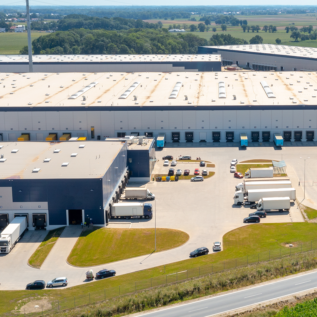 An aerial shot shows a vast industrial complex with multiple warehouses and numerous trucks and cars parked in the surrounding paved areas, all set against a backdrop of green fields.