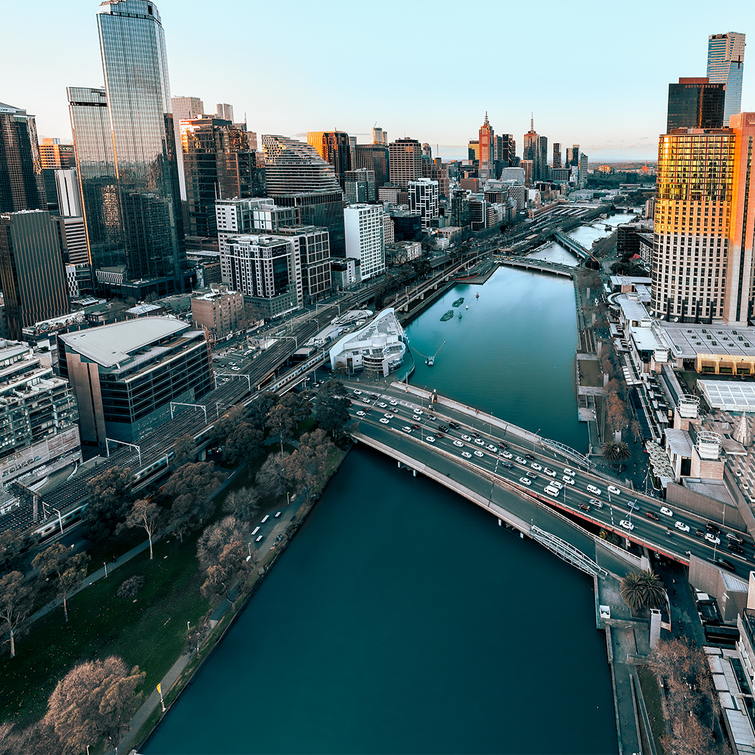 An aerial view of a bustling urban landscape featuring a river, multiple bridges with traffic, and numerous towering buildings under a bright sky.