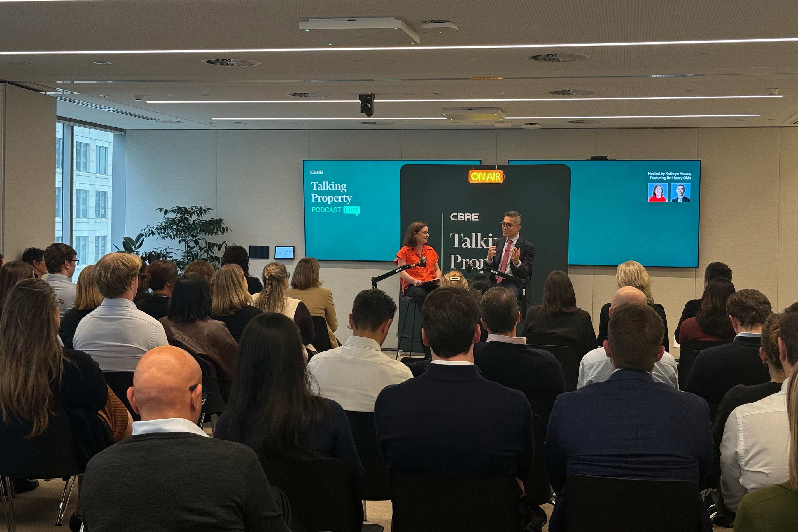 A diverse group of attendees face a stage where two speakers, Kathryn House and Henry Chin, sit in front of screens displaying "CBRE Talking Property Podcast Live" and an "ON AIR" sign.