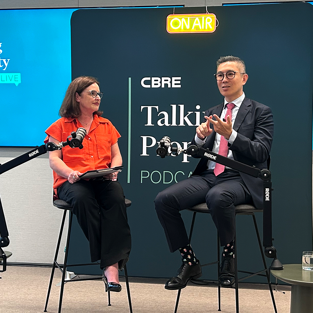 Two presenters, Kathryn House and Henry Chin sit on high stools with microphones, recording a podcast in front of a dark green banner that reads "CBRE Talking Property Podcast" with an "ON AIR" sign above them.