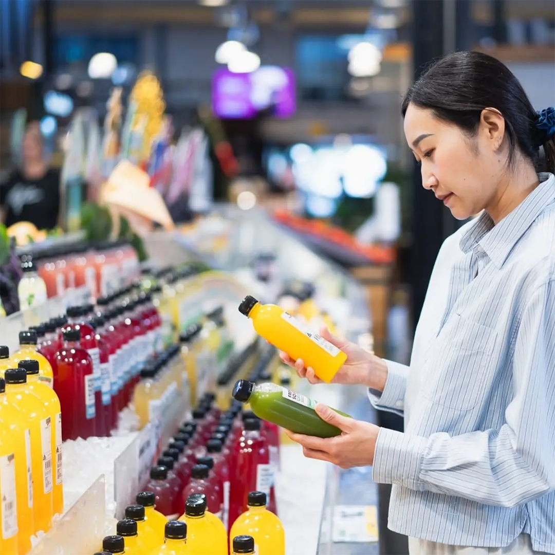 A woman in a light blue striped shirt holds a yellow bottle of juice in one hand and a green bottle in the other, examining them in a brightly lit supermarket aisle.